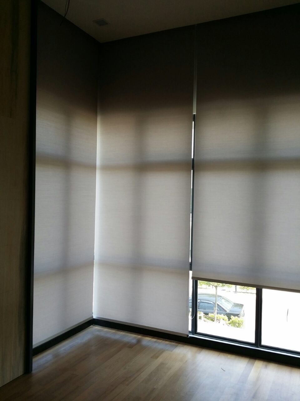 Light sunscreen roller blinds wrapping a corner of two windows, diffusing bright daylight over a wood floor