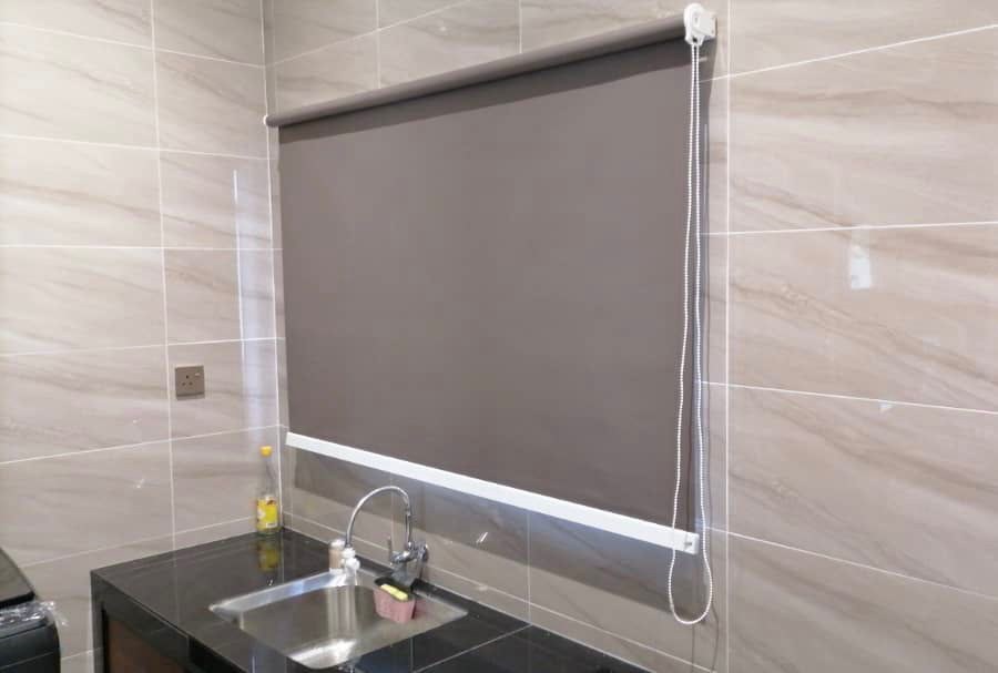 Grey blackout roller blind with a chain control fitted over a kitchen window above the sink, against marble-look tiles