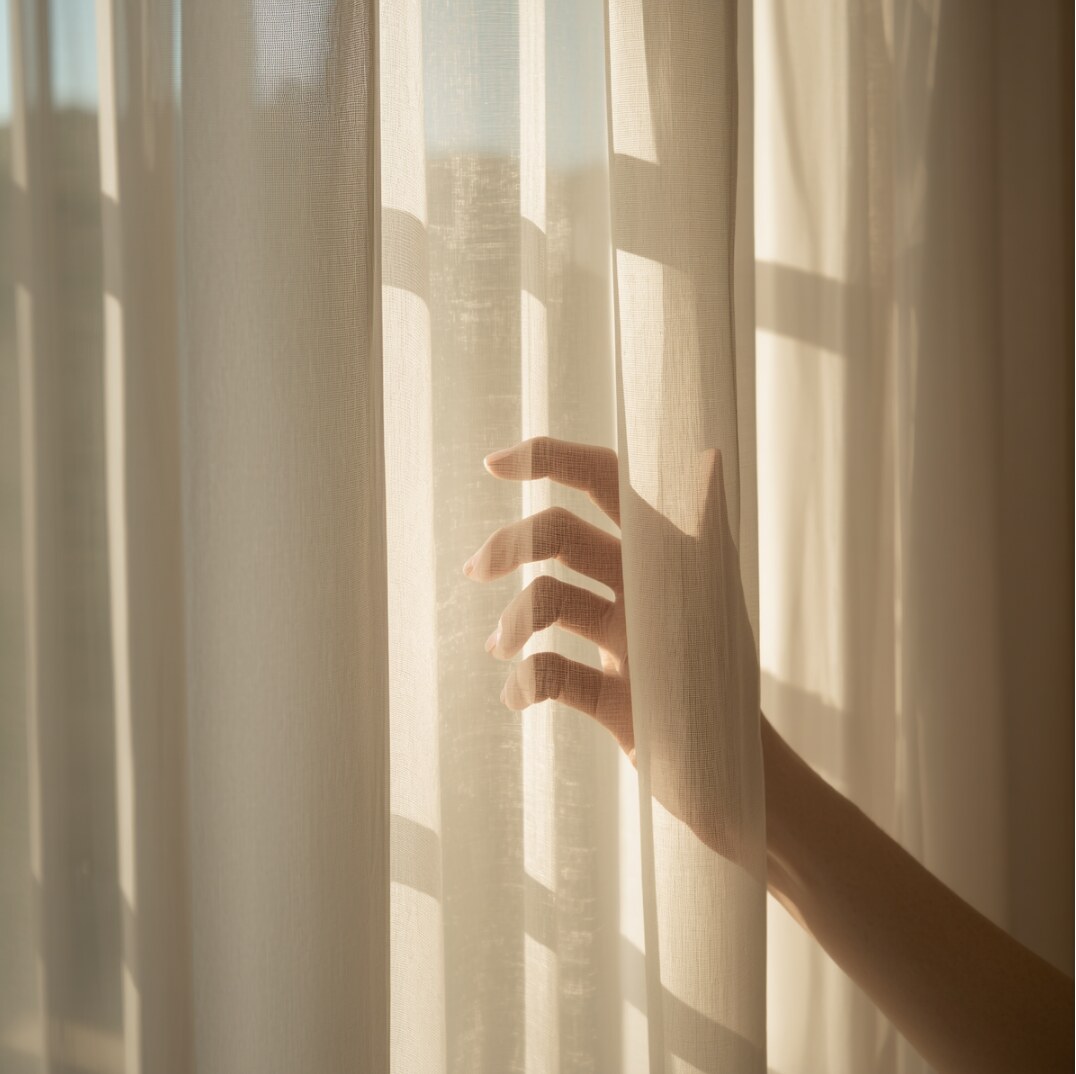 A hand parting a sheer oatmeal linen curtain in warm afternoon light